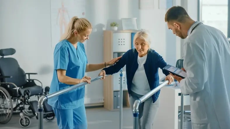 Rehabilitation nurse helps disabled senior walk using parallel bars