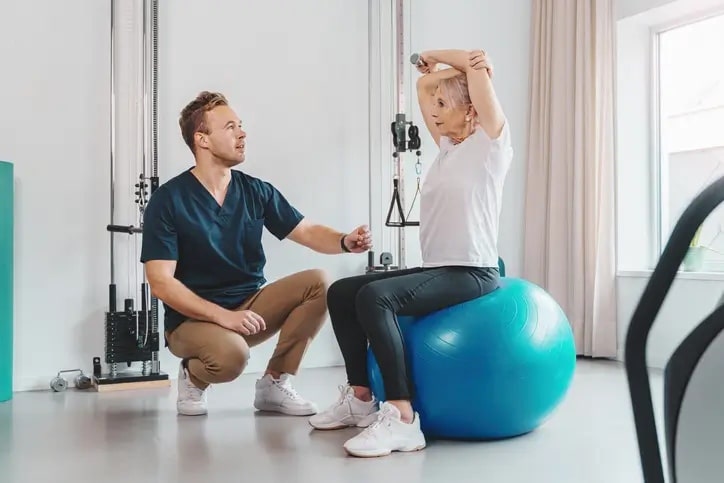 Senior woman sitting on pilates ball while nurse instructs physical therapy routine